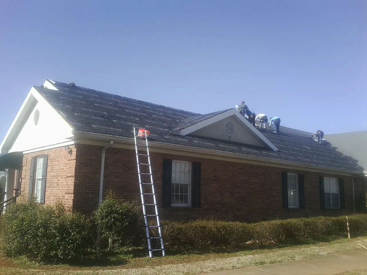 Crew tearing shingles off a brick ranch roof with a ladder set — Emergency Roof Repair work by our crew in Ardenvoir