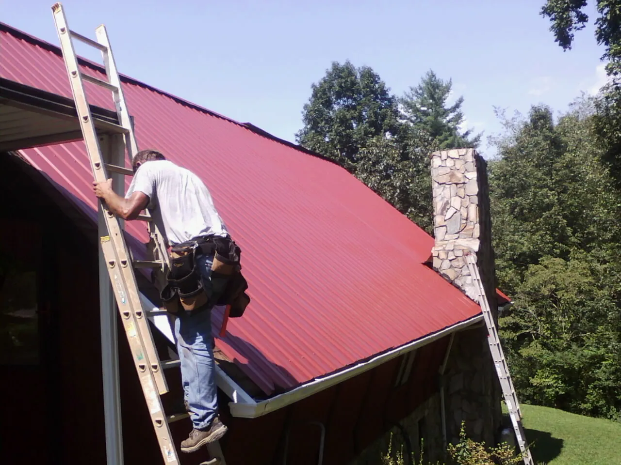 Roofer on a ladder beside a red metal roof and stone chimney — Soffit & Fascia Repair by Roofing Brothers in Ardenvoir